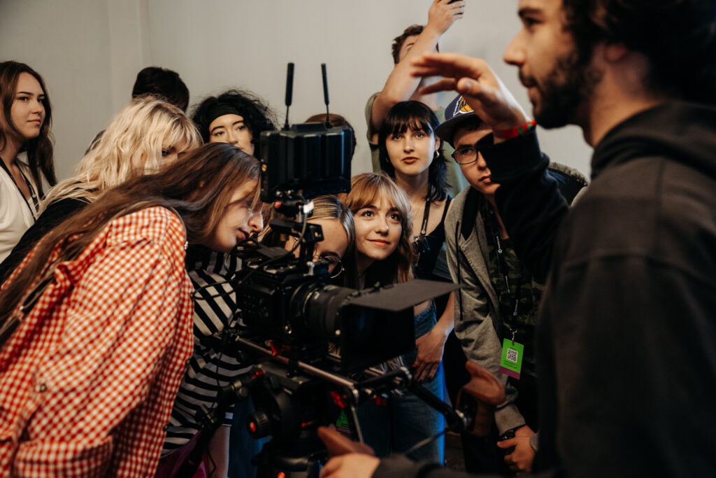 Osoby uczestniczące w warsztatach filmowych skupione wokół kamery podczas zajęć praktycznych.
